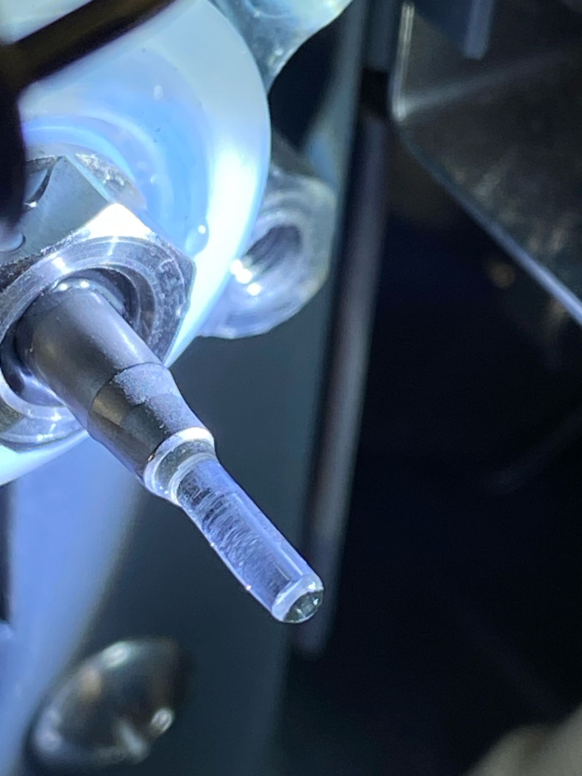 A sapphire plunger still mounted in the pump head, lit from the side against a dark background. A dull, frosted band runs along the length of the sapphire instead of a clear polished surface, and pale crystalline deposit sits on the metal collar behind it at the seal seat.